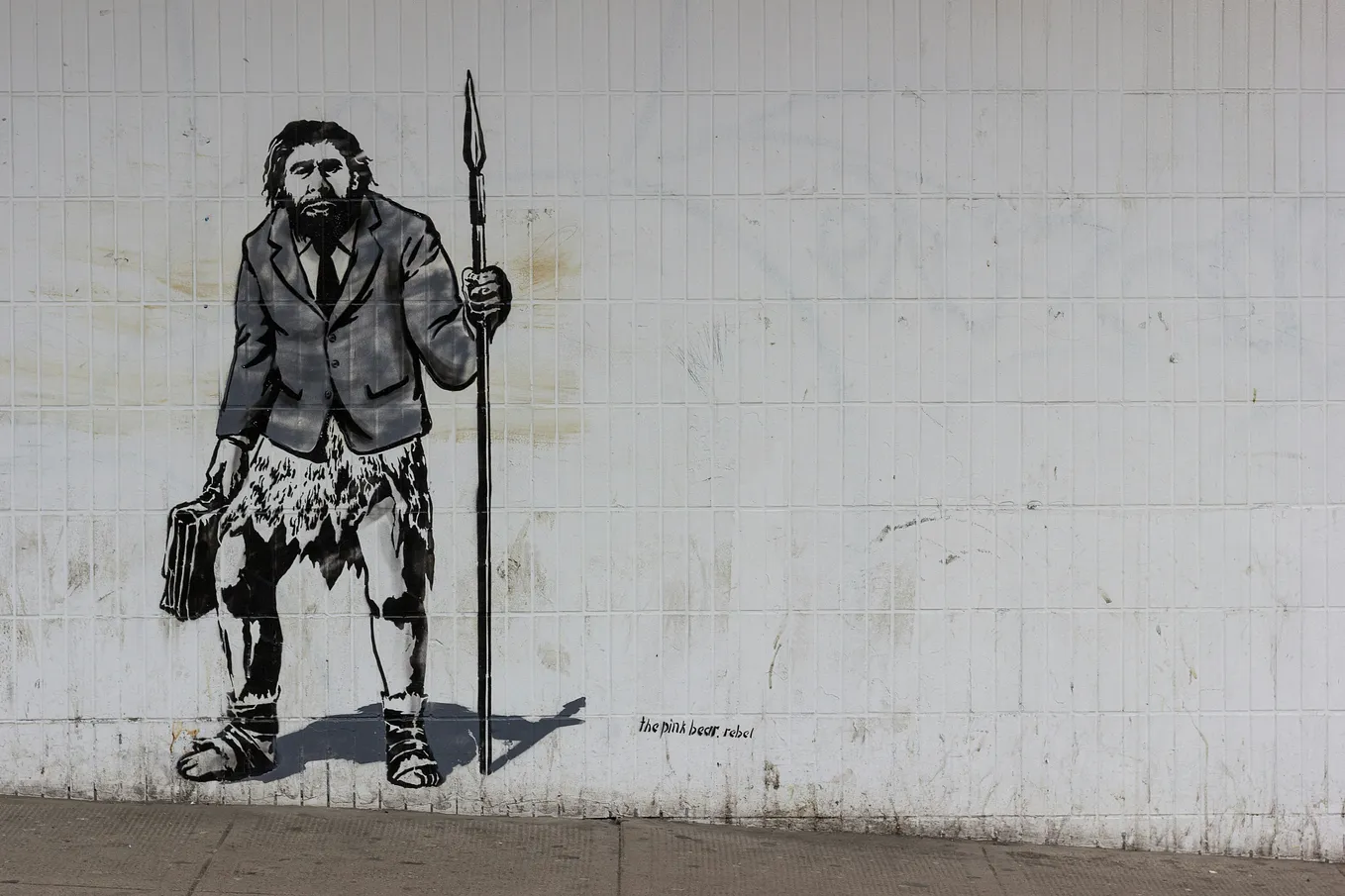 Street art mural depicting a caveman dressed in a modern business suit, holding a briefcase in one hand and a spear in the other, against a tiled wall background. The artwork is signed ‘the pink bear. rebel.’