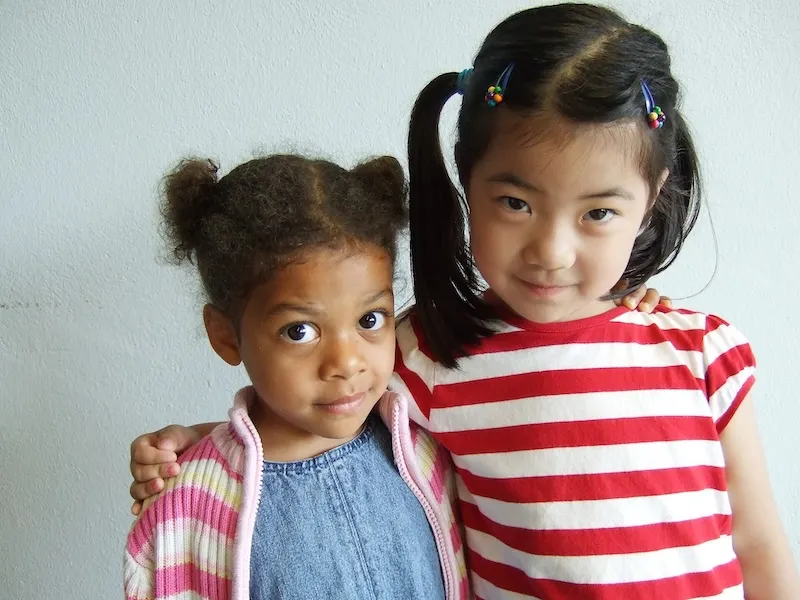 A Black child and Asian child facing the camera in a partial embrace against an off-white background. The Black child is a little girl with pigtails, wearing a blue shirt with a pink-and-white stripped cardigan. The taller Asian child is also wearing pigtails and is wearing a red-and-white stripped shirt.