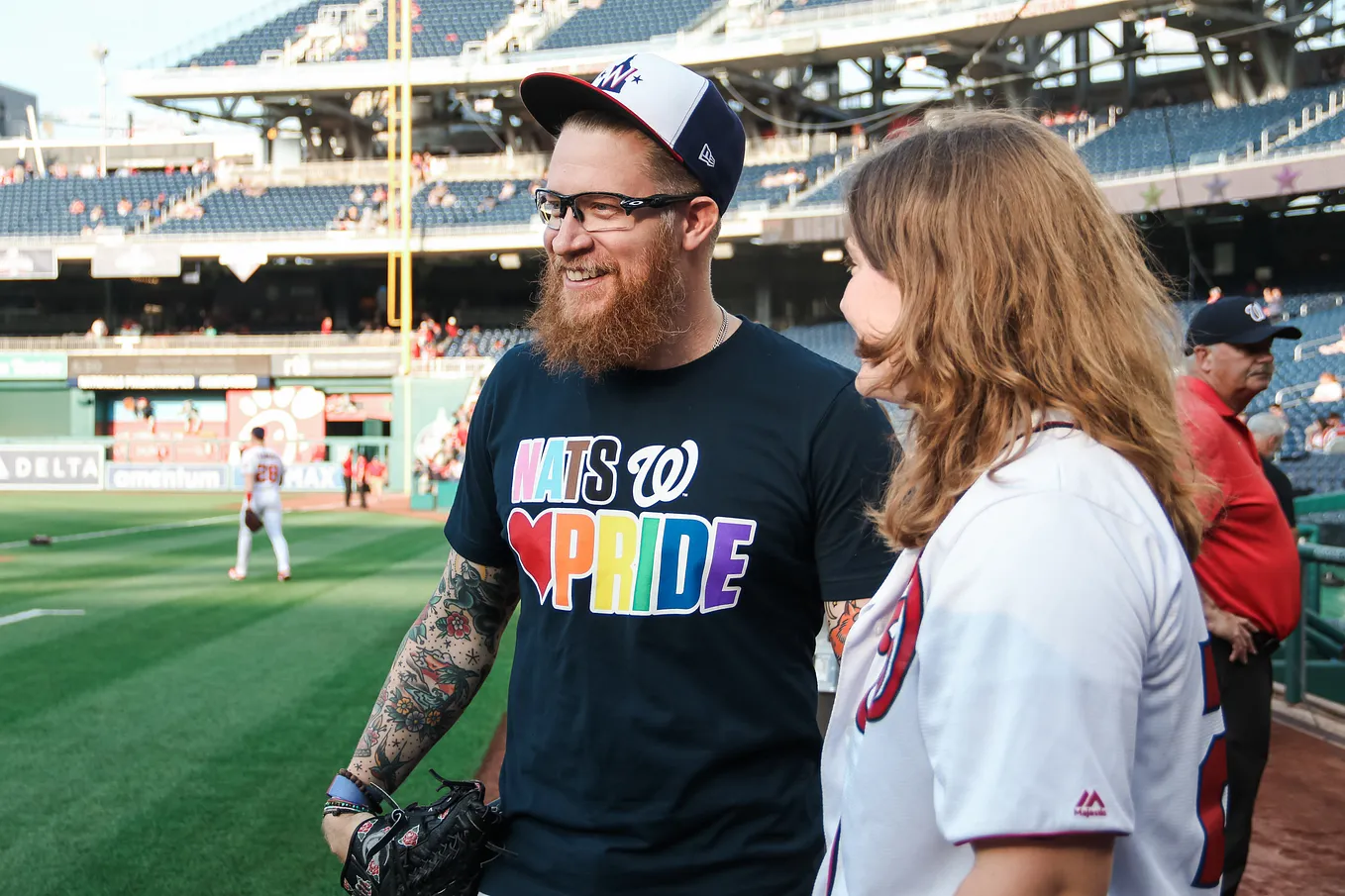 Sean Doolittle (center, with hat, beard and glasses) dons a “Nats (heart) pride” shirt and smiles as he speaks with a woman in the foreground on the right.