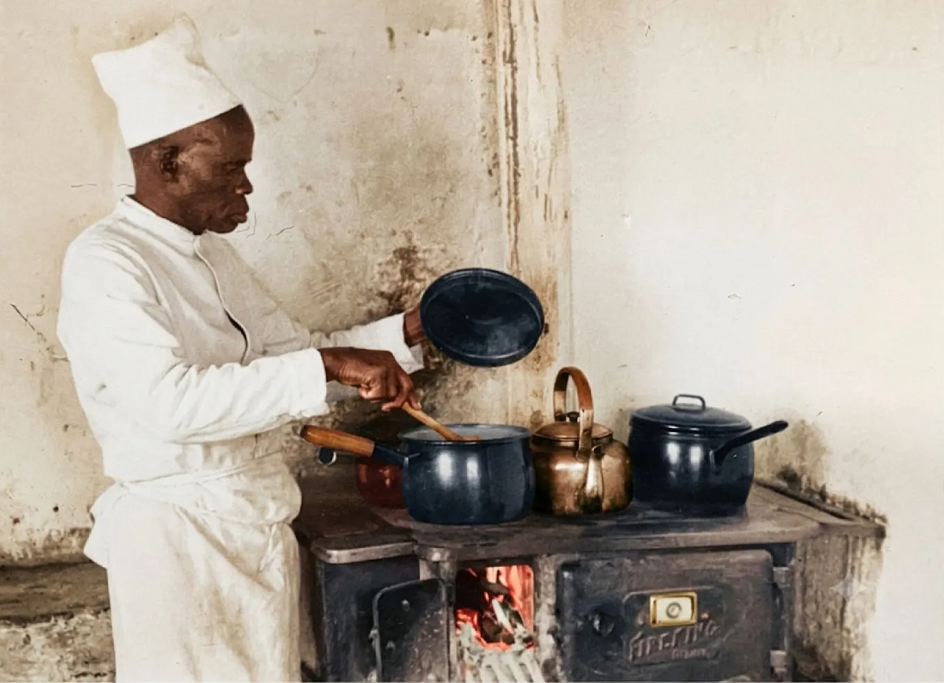 An African cook in a white apron and chef’s hat stirring a pot on a vintage wood-burning stove in a bush camp kitchen.