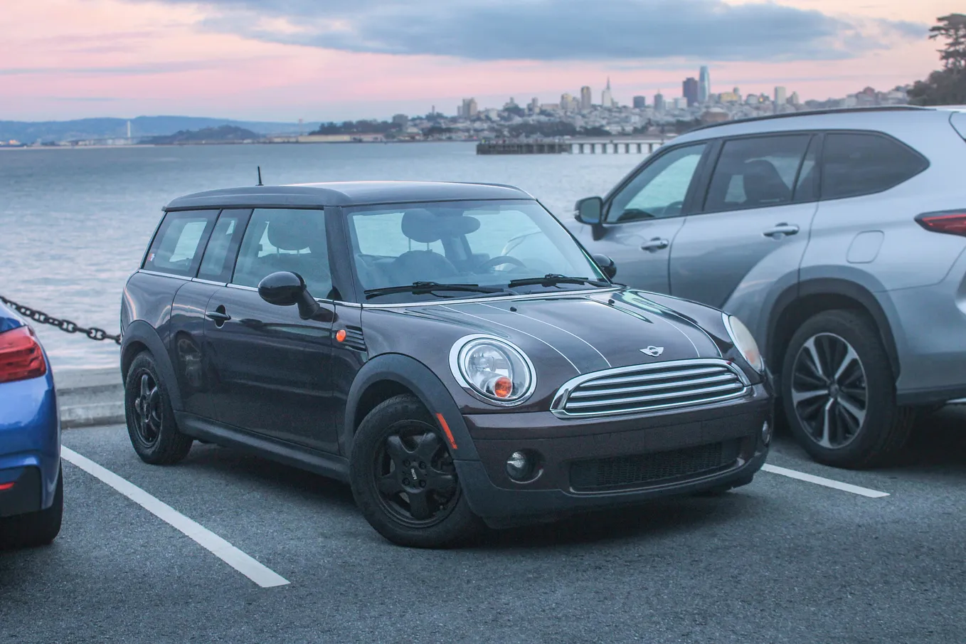 A Brown Mini Cooper Clubman with racing stripes and a sunset in the backround