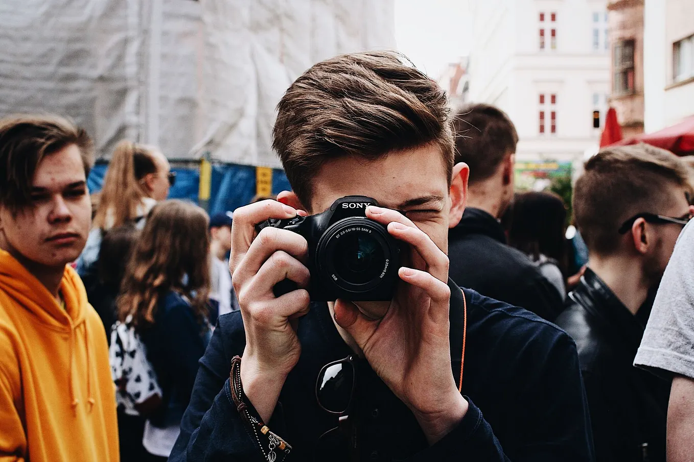 A young male photographer captures a bustling crowd with his camera outdoors.