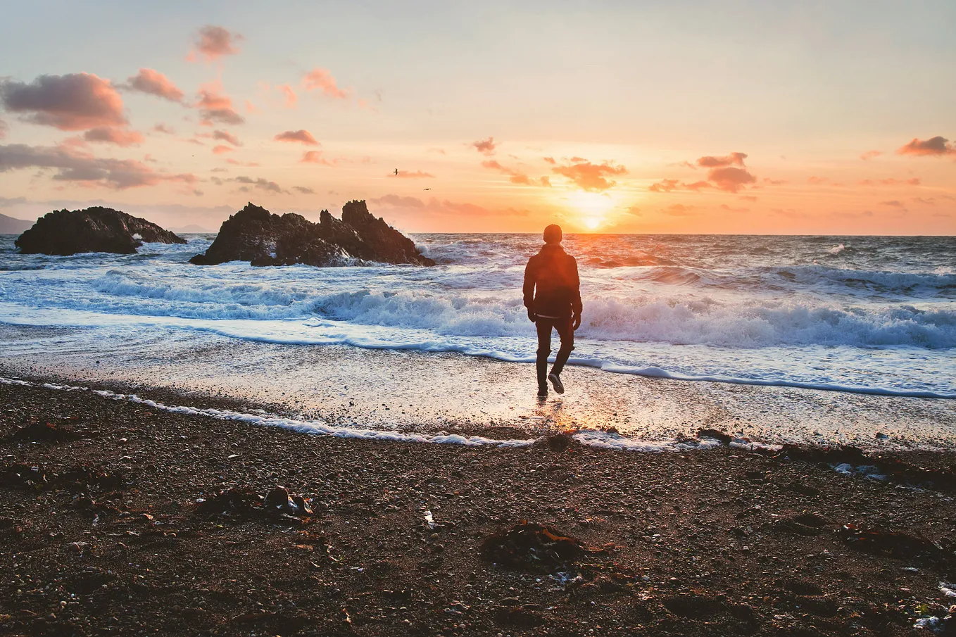 A person is running along a pebble beach towards the sea at sunset. The sun, low in the sky, casts a warm glow across the scene, highlighting the turbulent waves and rocky outcrops in the background. The sky is scattered with soft, orange-tinted clouds.