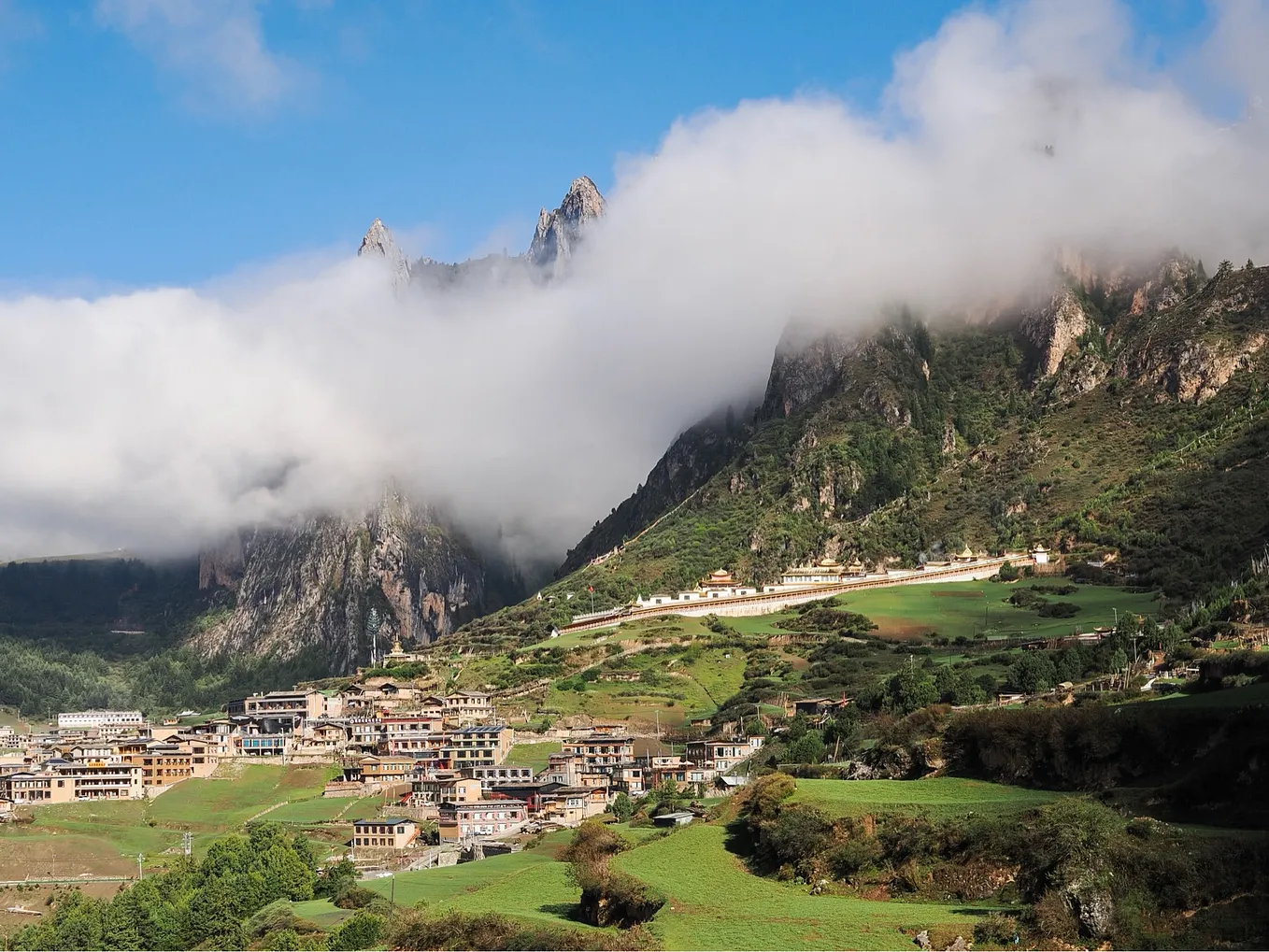 A village sits on a grassy slope below spiky mountains where low lying cloud wraps around the peaks