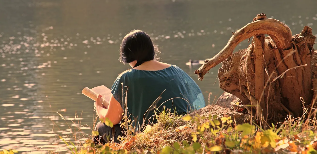 A young girl sitting on the bank of a lake, engrossed in a book.