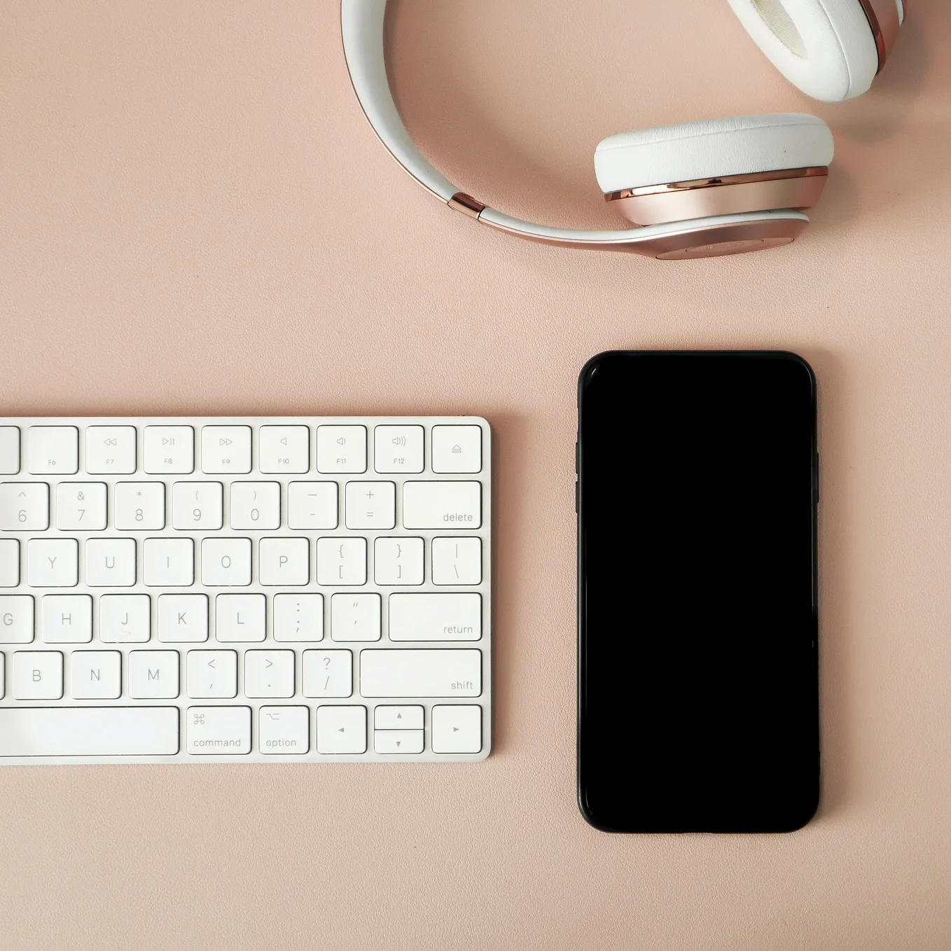 A beige coloured desk. On top sits a white computer keyboard, a pair of rose-gold headphones and a mobile phone switched off.