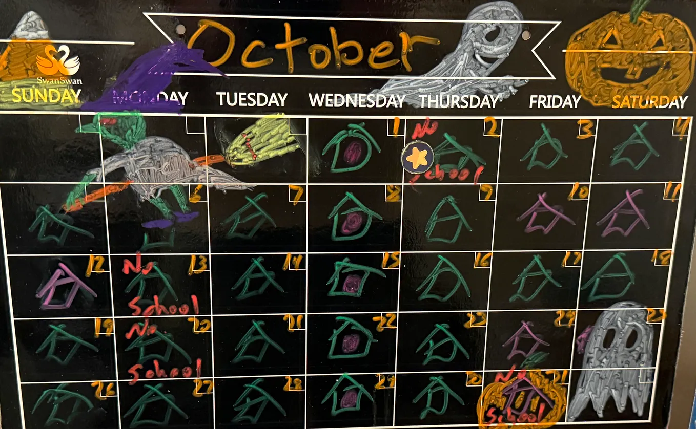 A black wall calendar with the month drawn by hand, a small house in each day square in green or purple, a ghost and a jack-o’-lantern across the top, and a star magnet on the first.