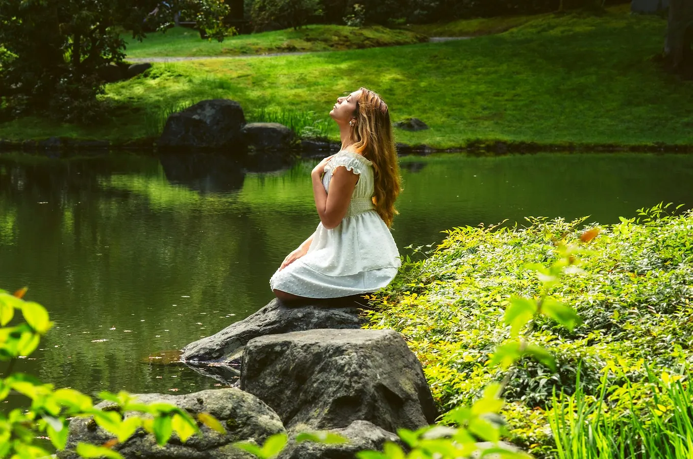 A woman in a white dress holds a hand to her heart and closes her eyes while sitting on a rock beside a river surrounded by green grass and foliage.