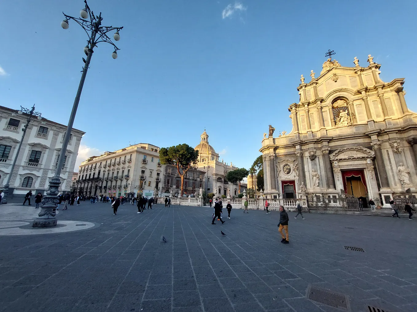 Catania cathedral lit up by low afternoon sun. The sky is blue, and a few people walk through the square. The statues on the church stand out above the dark piazza.