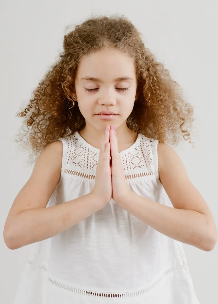 Photo of a young girl with curly hair, eyes closed and hands in “namaste” pose