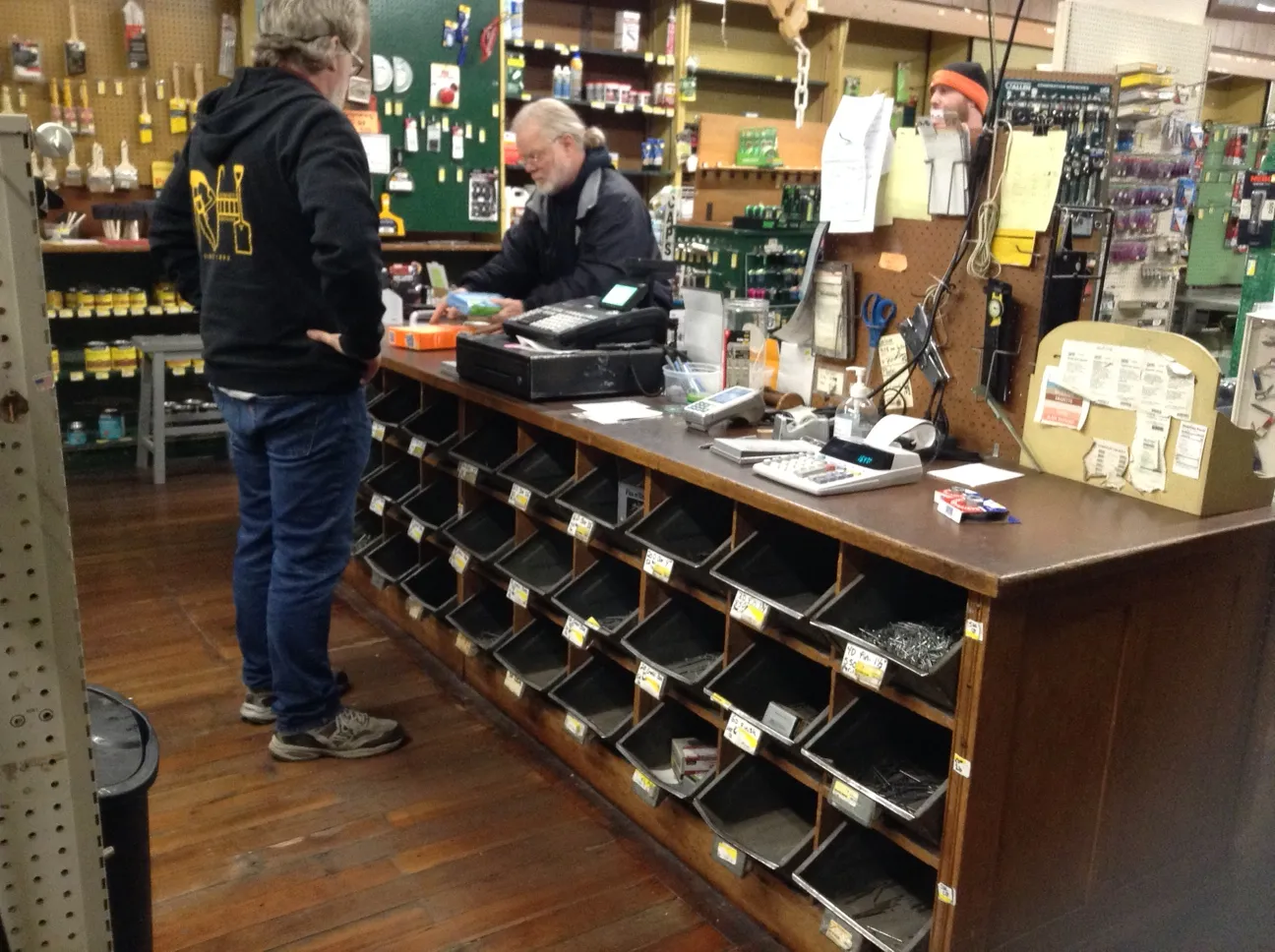 An employee standing behind and old wooden counter with mental bins. A customer on the other side completes a purchase. Walls in the background are covered with paitn brushes, stains, and wrneches.