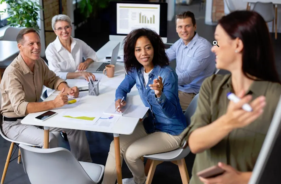 A photo of a woman standing at a flip chart with a marker, speaking to a group of four other people around a table.