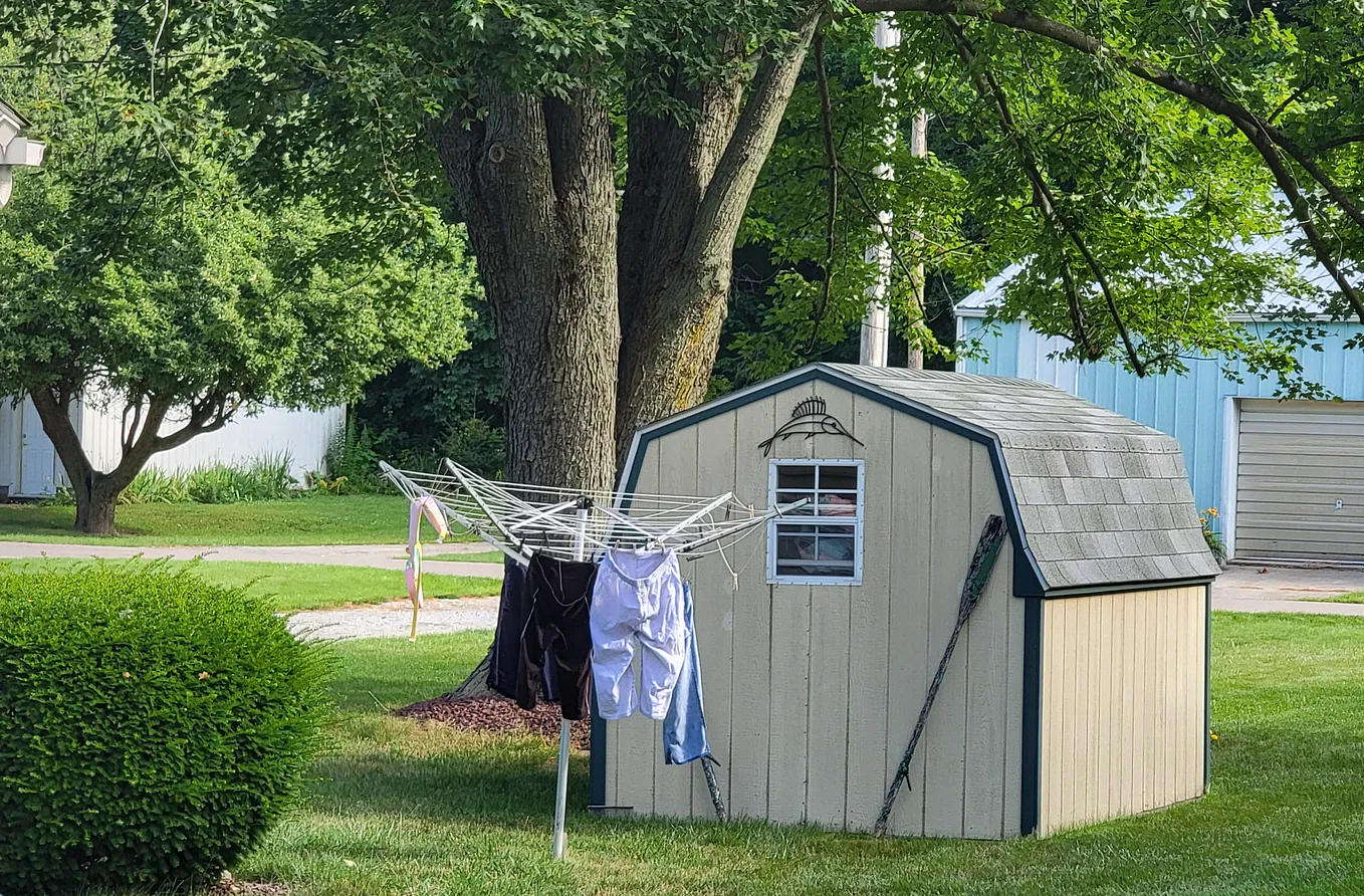 A small, tan garden shed with a folding drying rack in front of it, with some clothes drying.