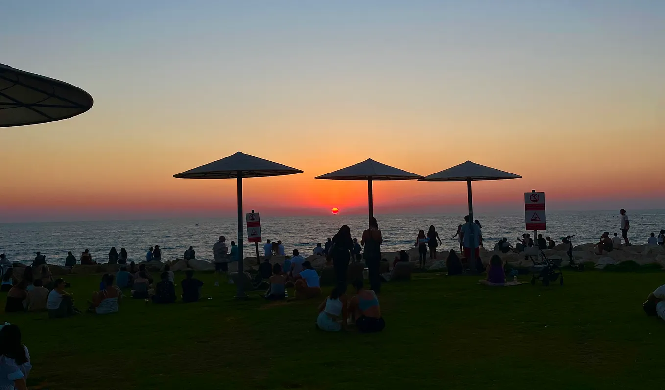 The sun sets over HaTuppim Beach in Tel Aviv, Israel — umbrellas and some beach goers are in silhouette against the setting sun.