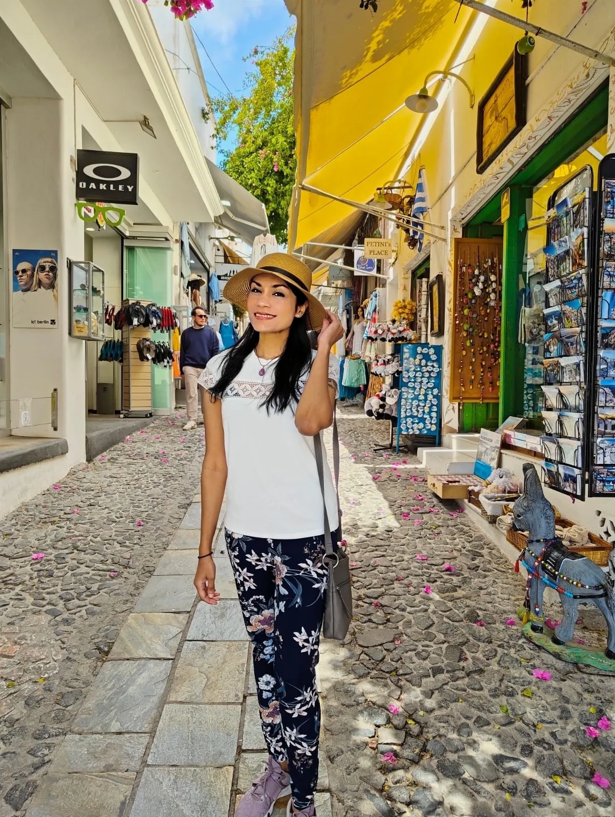 A West Indian girl wearing a straw hat, a white top, and floral pants is smiling for the camera. The picture was taken in Santorini, Greece. A grey handbag hangs around her shoulder.