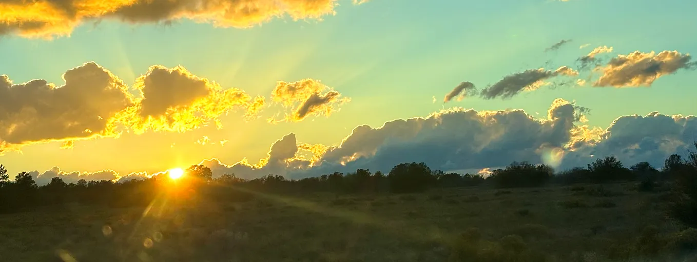 A bluish-aqua sunset with bright yellow-gold filling and lining the clouds and rays stretching into a shadowed foreground desert landscape.