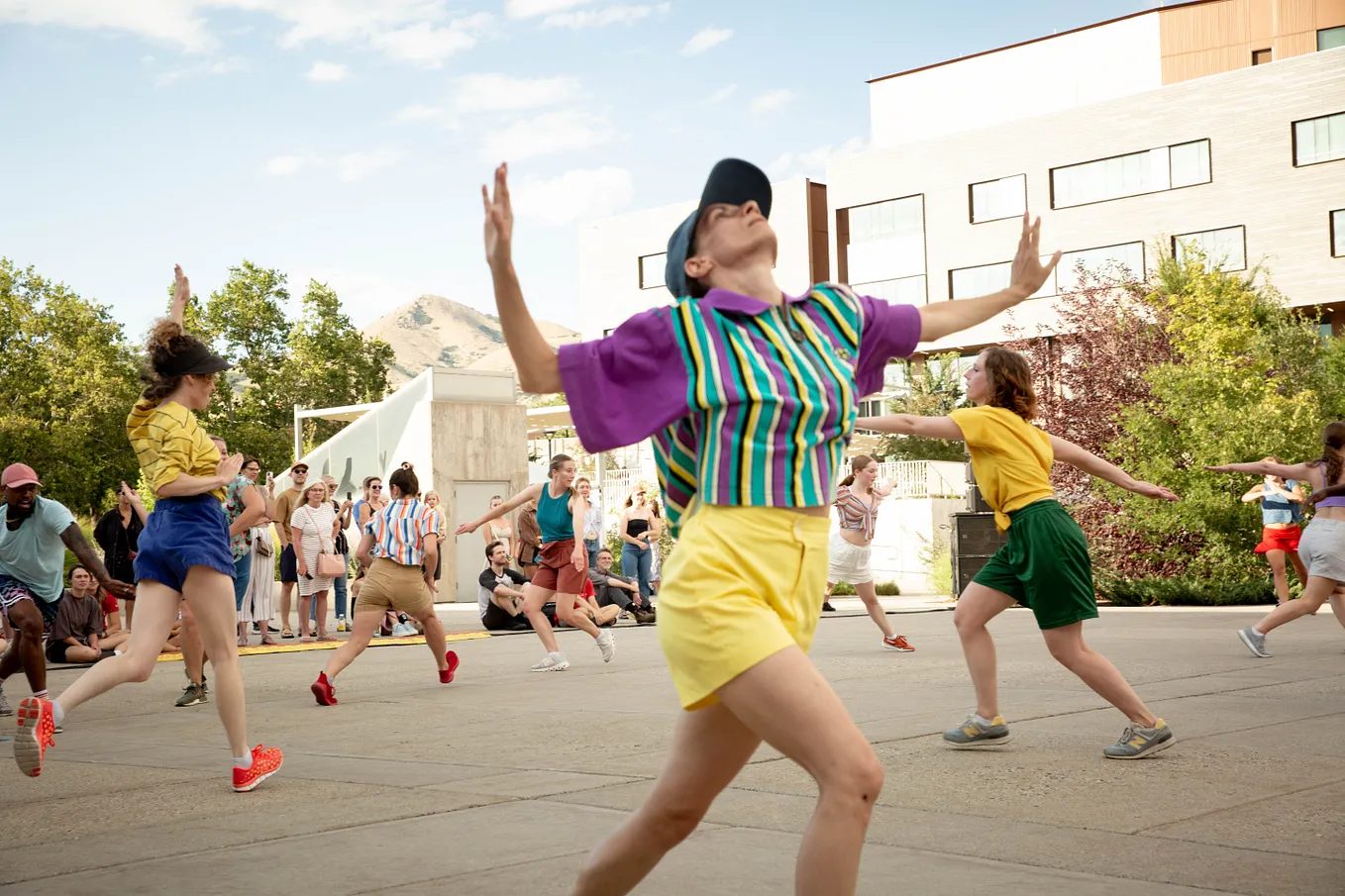 A group of dancers performing outdoors in a plaza. The daner in the foreground wears a striped shirt, baseball cap, and yellow shorts.