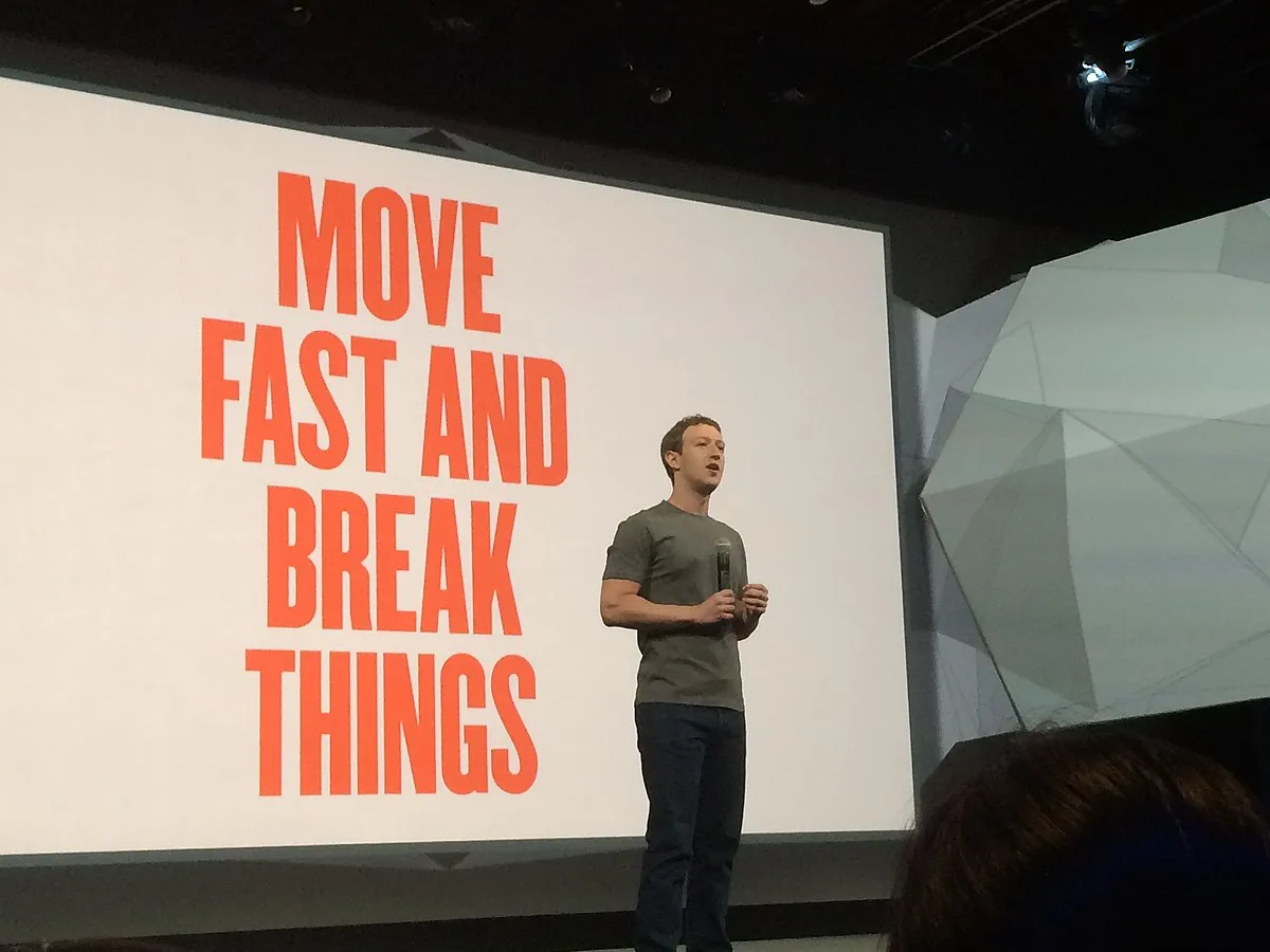 IMAGE: A yaoung Mark Zuckerberg standing in the scenario with the motto “Move Fast and Break Things” in red capital letters behind him