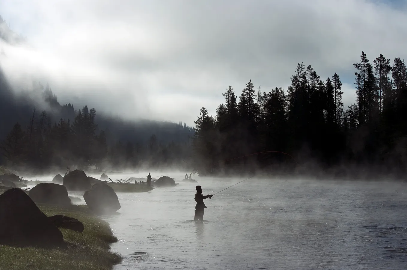 Black and white photo of a man fly fishing on a mist-covered river at sunrise.
