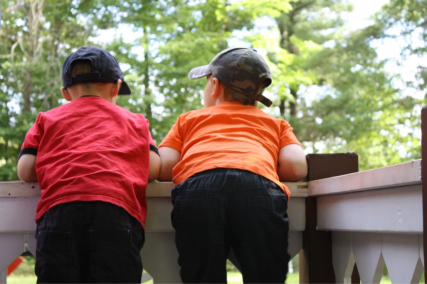 A photo taken from behind of two little boys in red and orange shirts and baseball caps leaning on a wooden railing facing a line of trees.