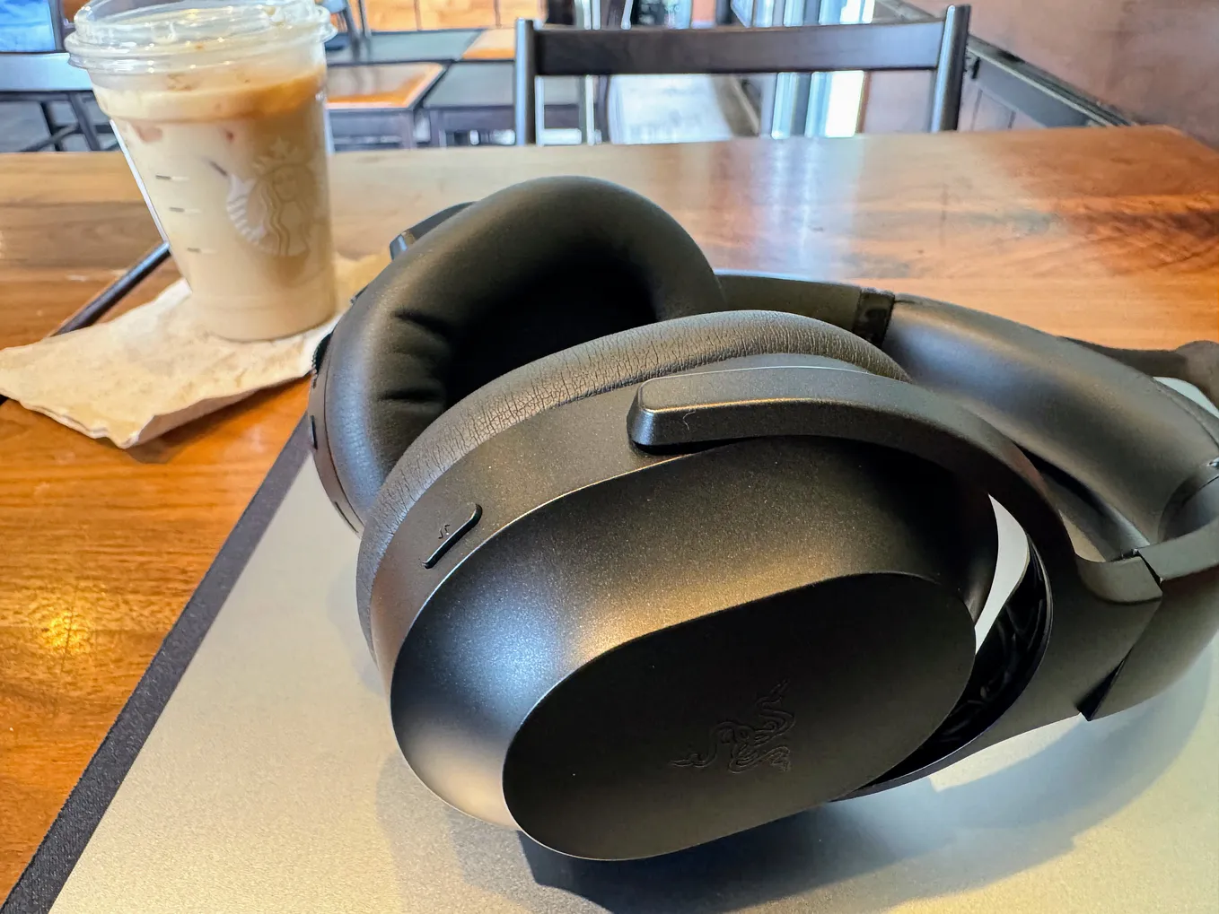 A Razer Barracuda Pro gaming headset lying on its side on a closed laptop on a table. A cup of iced coffee sits on the left side of the frame.