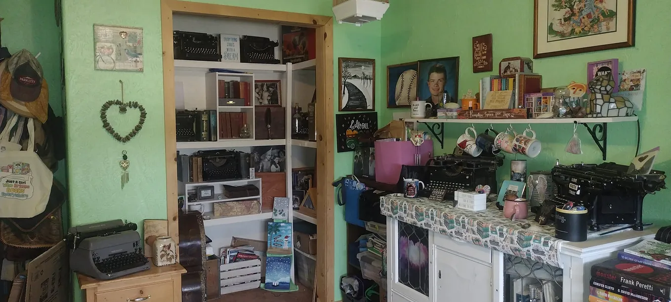 Home office with light green walls and a small end table on the left corner holding a typewriter, a white cabinet on the right holding two typewriters and several knick knacks and a closet displaying several typewriters and old books.