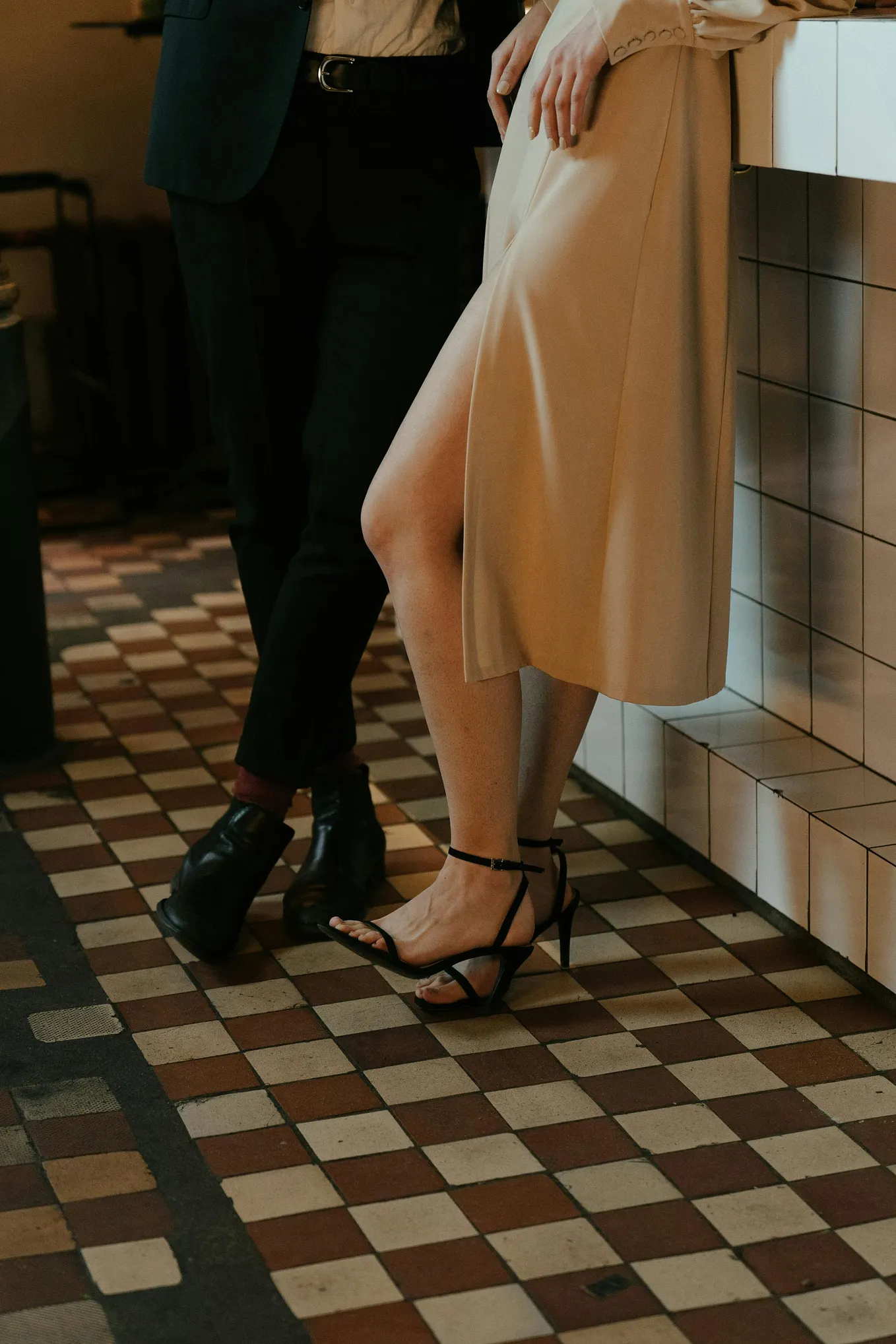 A man in black trousers and black leather shoes stands next to a woman in black strappy stillettos. They’re standing on a brown and white tiled floor leaning against a white tiled drinks bar, we can only see their bottom half.