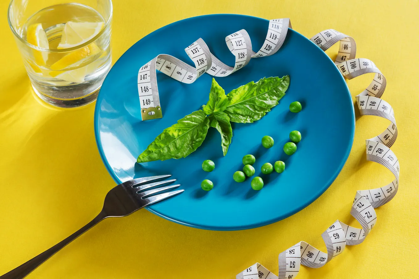 A blue plate on a yellow background with a minimalist arrangement: a few green peas, a large basil leaf and a fork. There is a rolled up tape measure around the plate. Next to it is a glass of water with lemon slices.