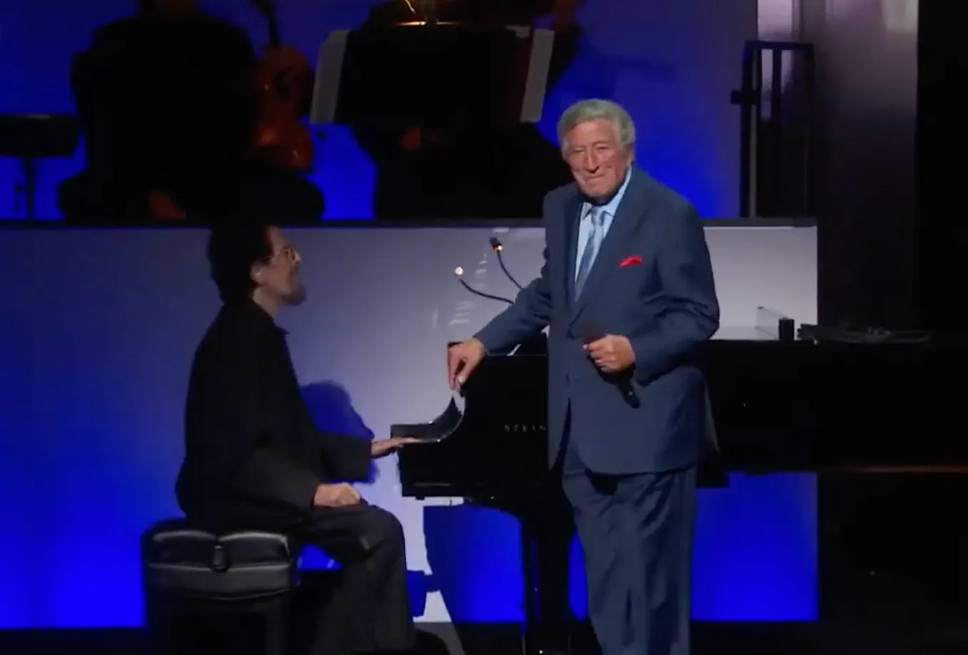 Tony Bennet, 89, on stage, dressed in a suit, leaning his right hand on a piano, microphone in his left hand.
