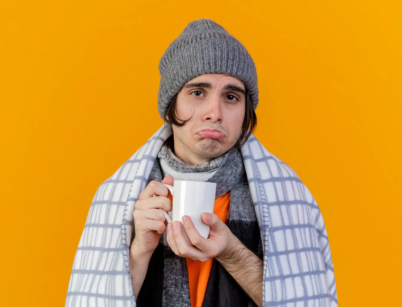 Photo of sad young ill man wearing a winter hat and scarf. He’s all wrapped up in a blanket, holding a mug of tea. Against a yellow background