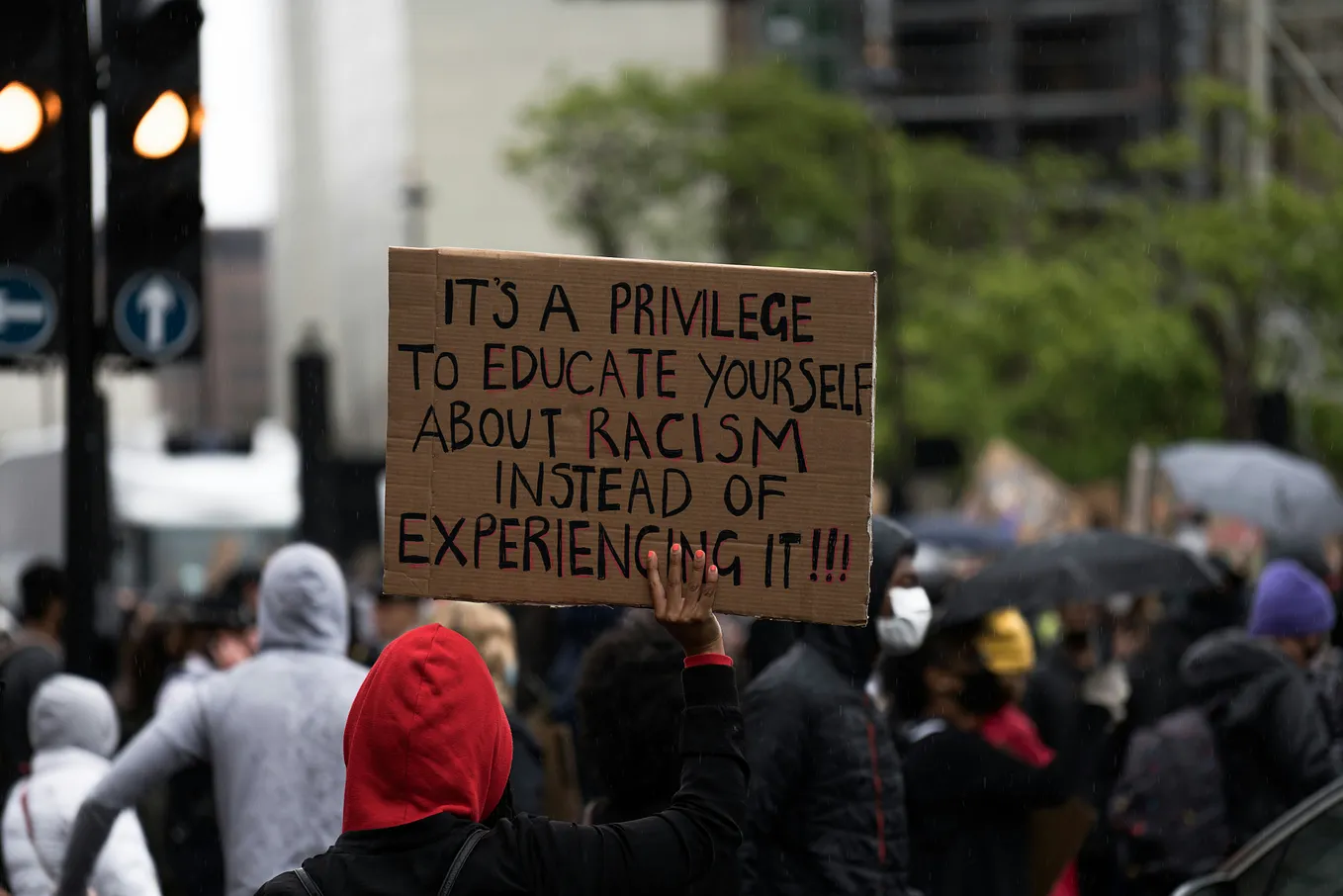 A person holding a sign at a protest that reads ‘It’s a privilege to educate yourself about racism instead of experiencing it!!!’ The person is wearing a red hoodie, and there are other people and buildings visible in the background.