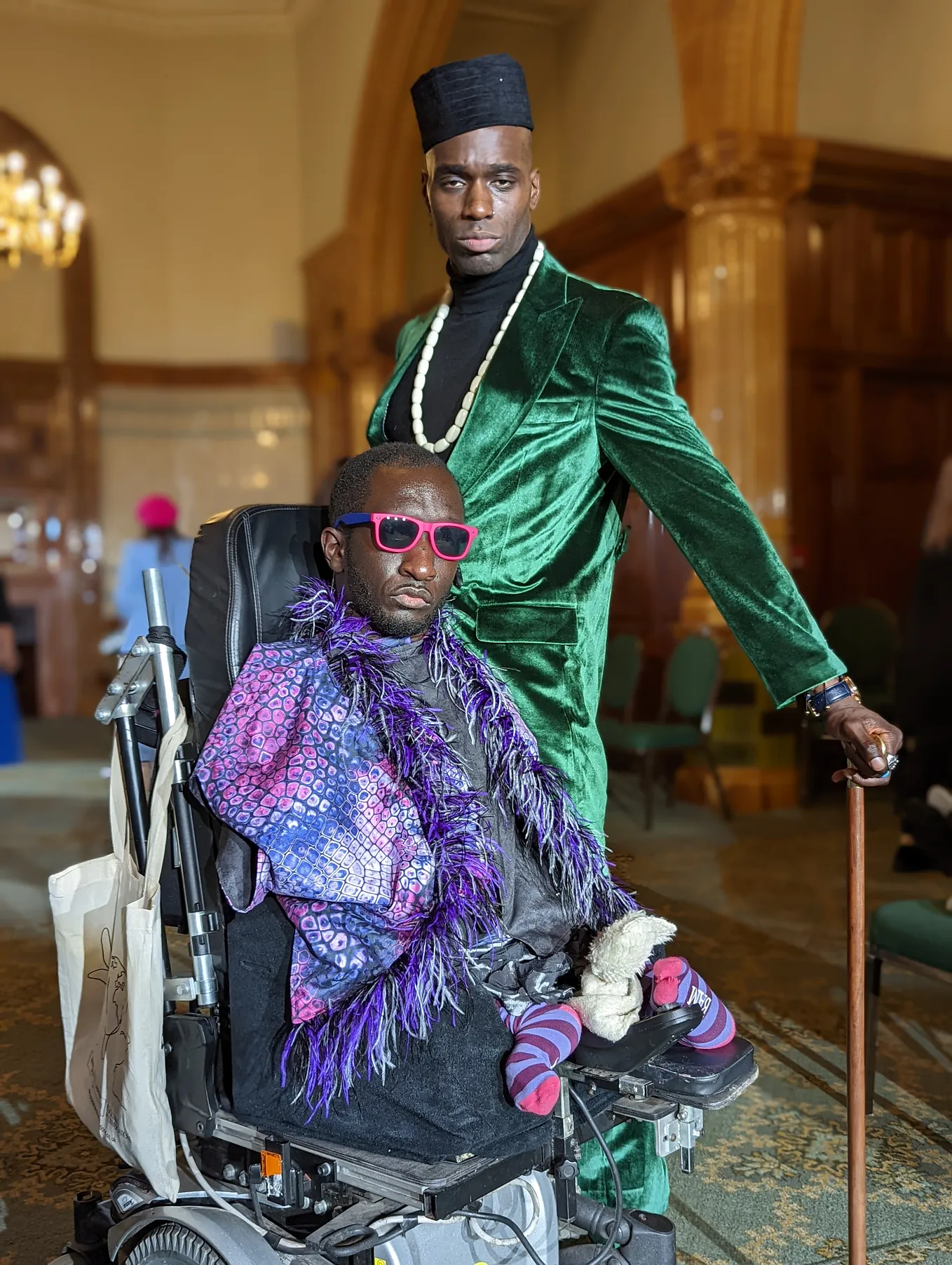 Two Black men dressed up, one standing and one seated in his wheelchair, are posing for the camera.