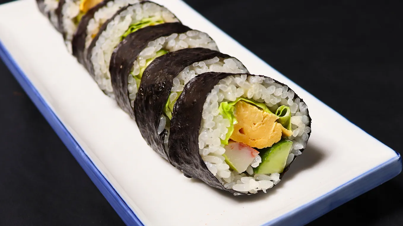 A close-up of a sliced sushi roll filled with rice, lettuce, cucumber, imitation crab, and tamagoyaki, wrapped in nori seaweed, served on a rectangular white plate.