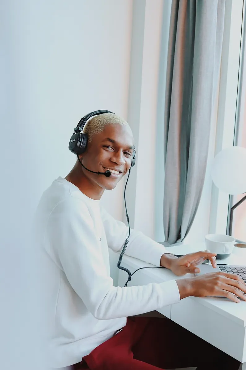 Image of young person, with close cropped hair, looking at camera and smiling brightly. He’s sitting at a laptop and is wearing a headset with a mic.