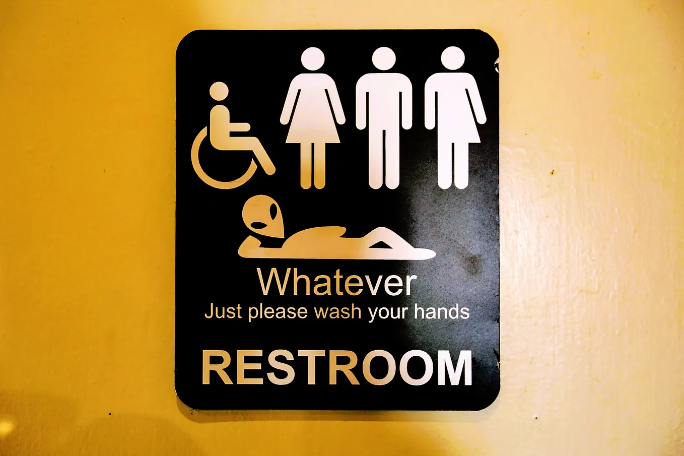A toilet sign with a wheelchair, a man, woman, unisex and a little green alien man. “Whatever, just please wash your hands” is the sign caption.
