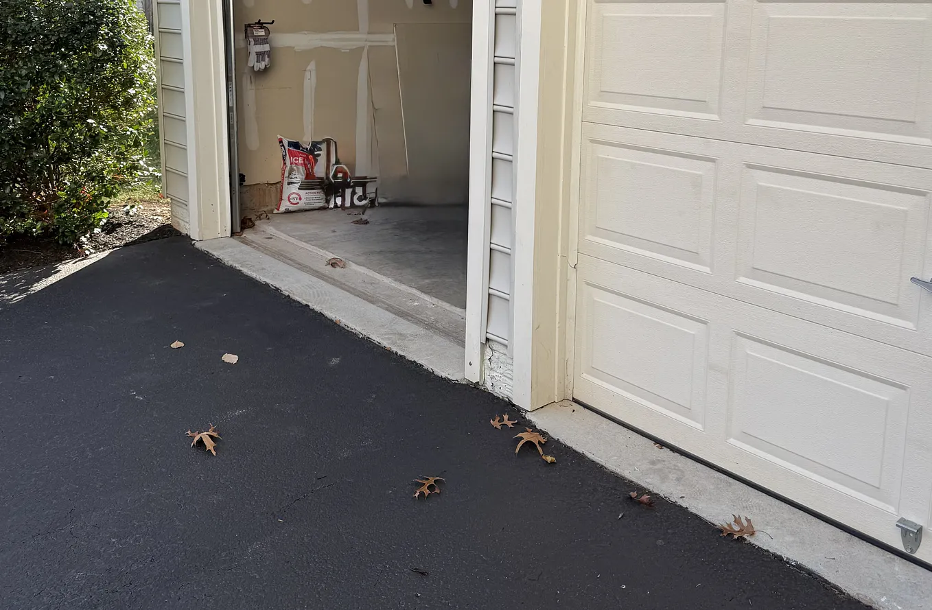 An open single garage bay seen from the driveway, dark inside, dead leaves scattered on fresh blacktop.