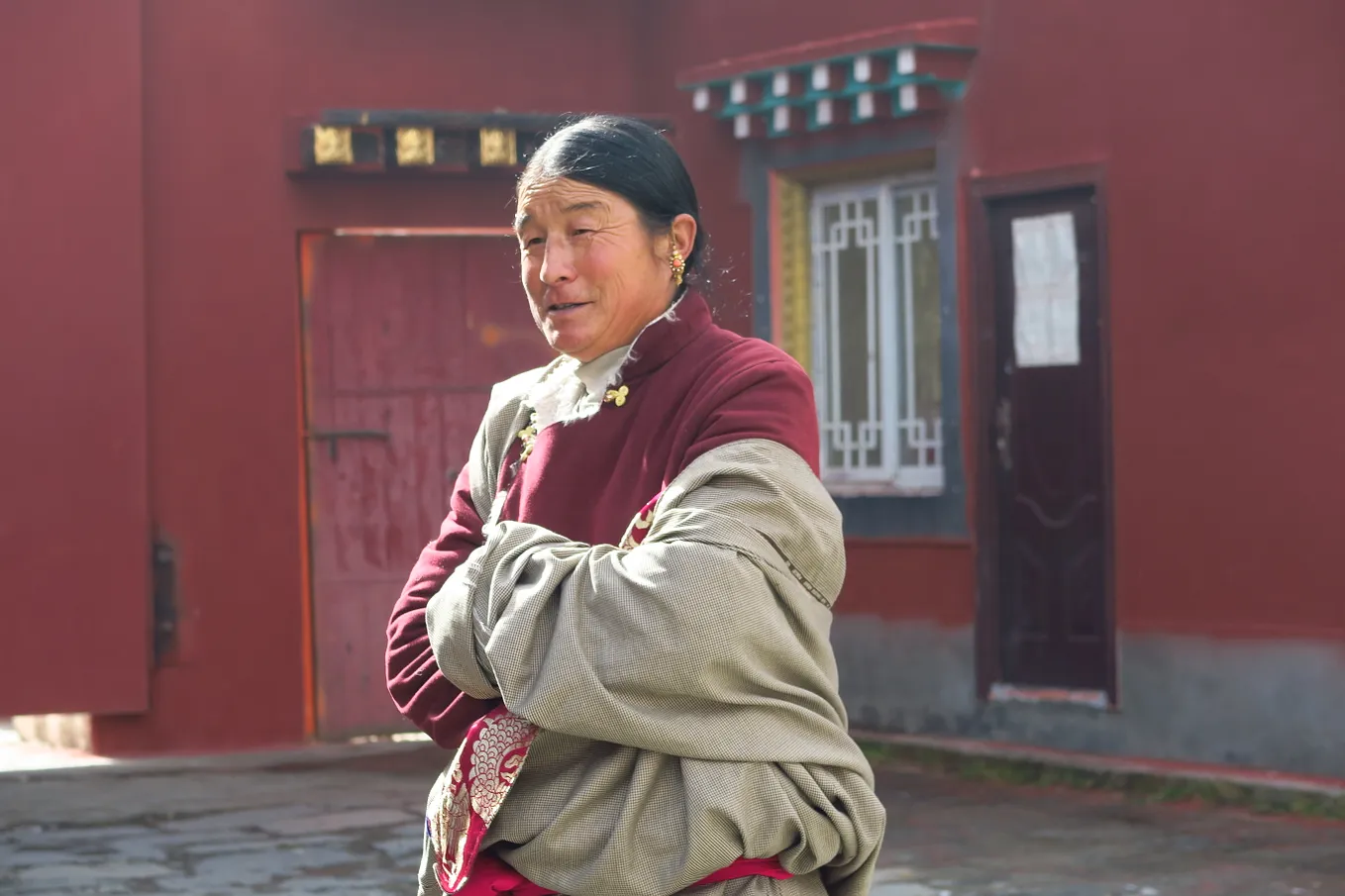 A tibetian woman standing against a red wall building. A sun is shining on her face and she is wearing a smile.