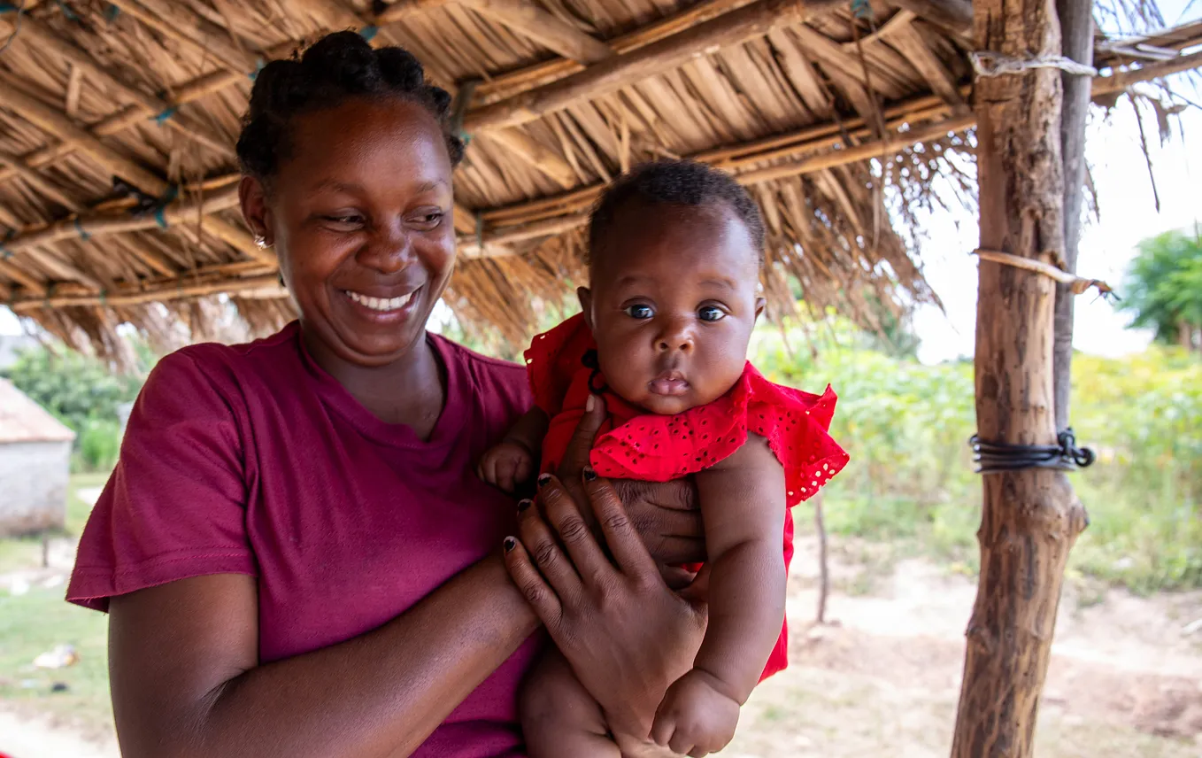 A mother gazes joyfully at her daughter who she is holding in her arms. The baby is staring intently into the camera capturing her image.