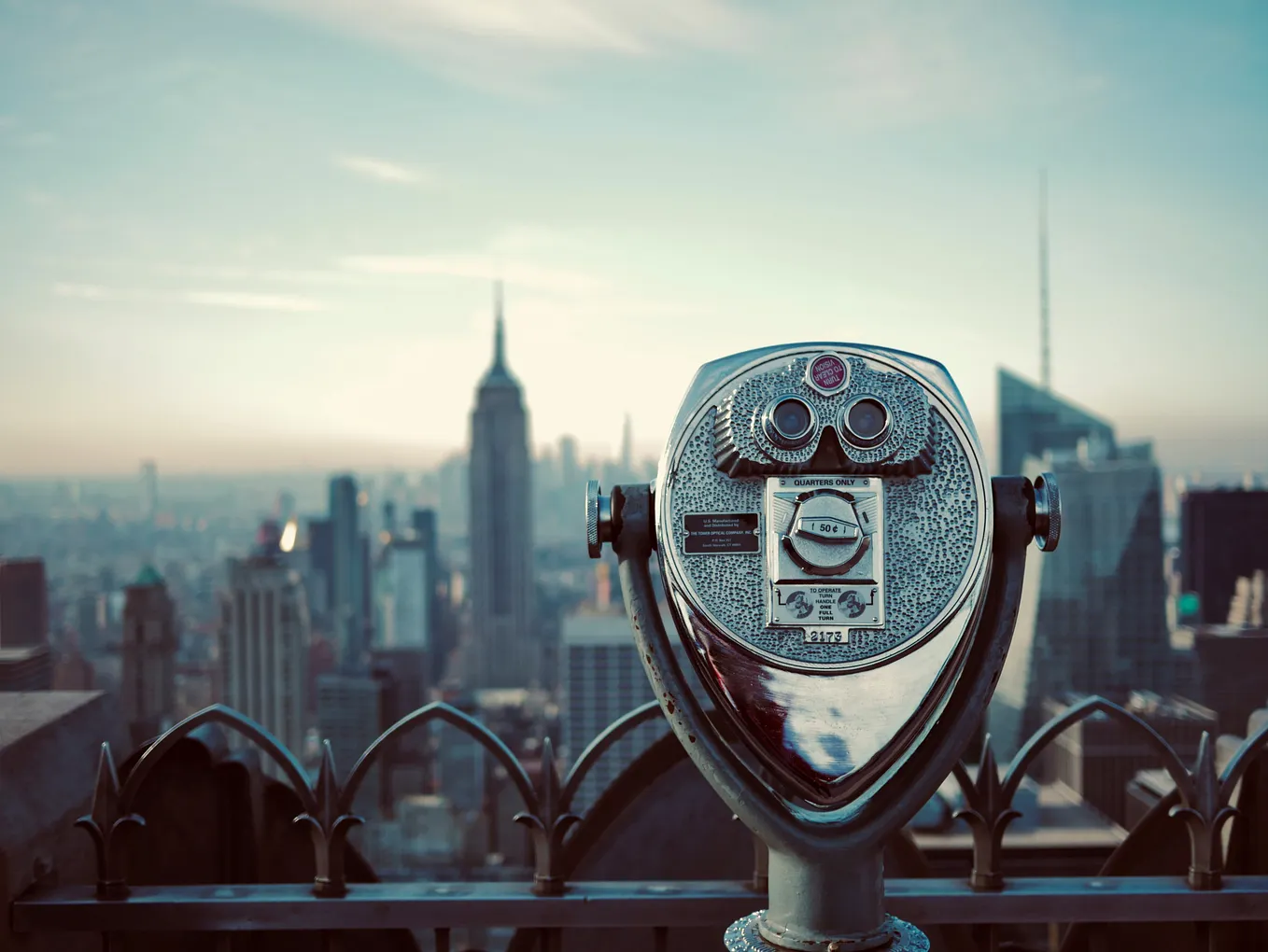 A vintage coin-operated telescope viewer on an observation deck overlooking the New York City skyline, with the Empire State Building prominently visible in the background among other skyscrapers under a hazy sky.
