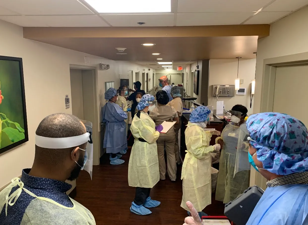 Healthcare professionals in protective gear, including gowns, face shields, and masks, gather in a hospital corridor for a briefing or meeting, likely related to infection control or patient care.