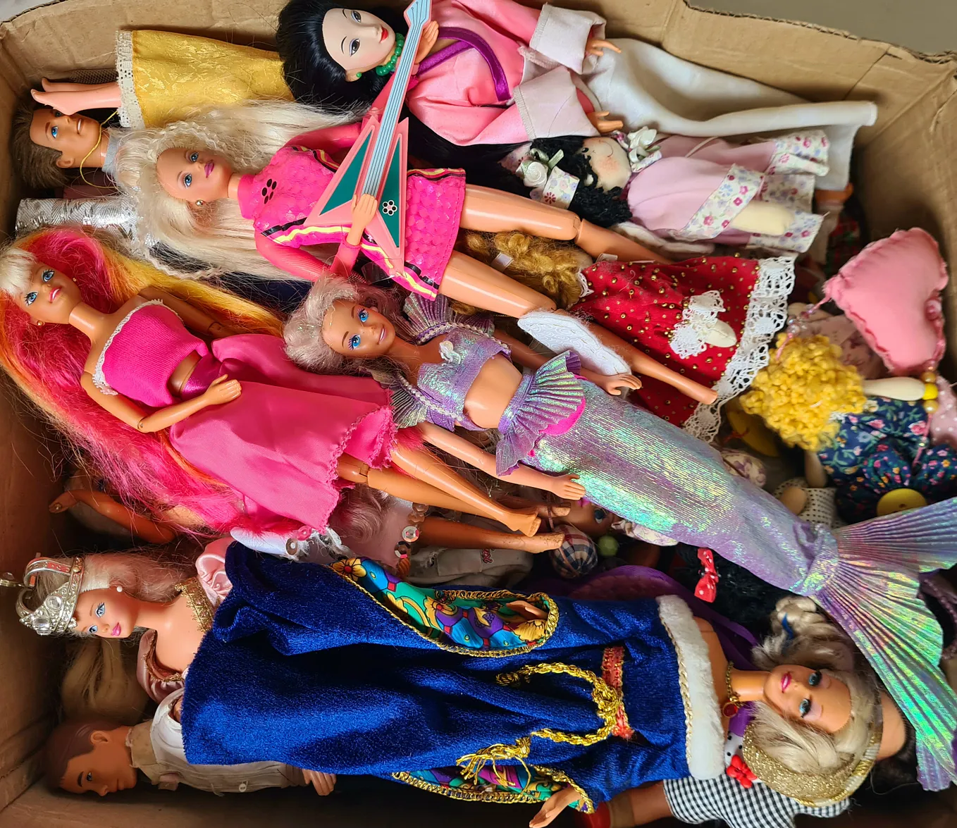 The image features a cardboard box full of Barbie dolls and a few Ken dolls. In the foreground, there’s a mermaid Barbie with a purple fishtail, a rockstar Barbie holding her pink guitar, a Hawaiian Barbie not in her original clothes, a princess Barbie and a Mulan Barbie.