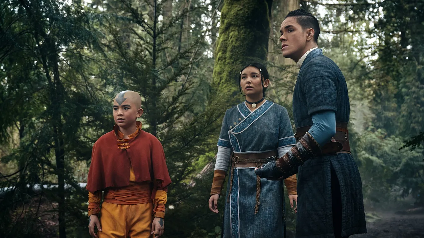 A trio of young people stand in the forest, each looking out of frame, appearing wary of something.