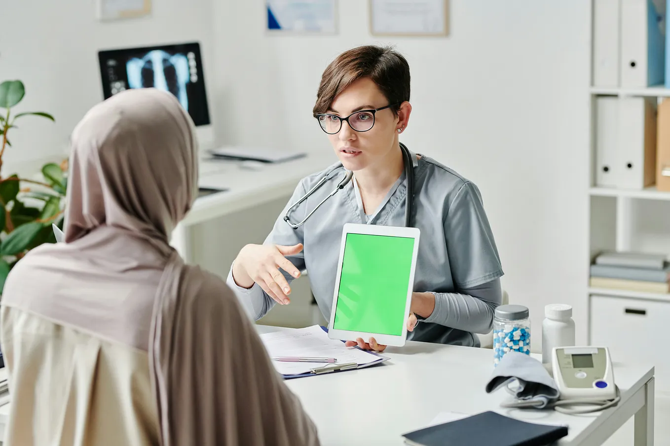 A healthcare professional holding a tablet with a green screen, explaining something to a patient wearing a hijab in a medical office setting.