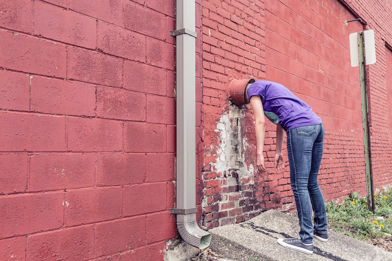 Woman standing with head stuck in a pipe coming out of the side of a building.