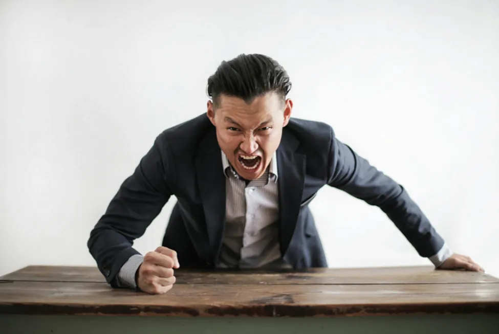 A photo of an angry man in a suit pounding his fist on a desk.