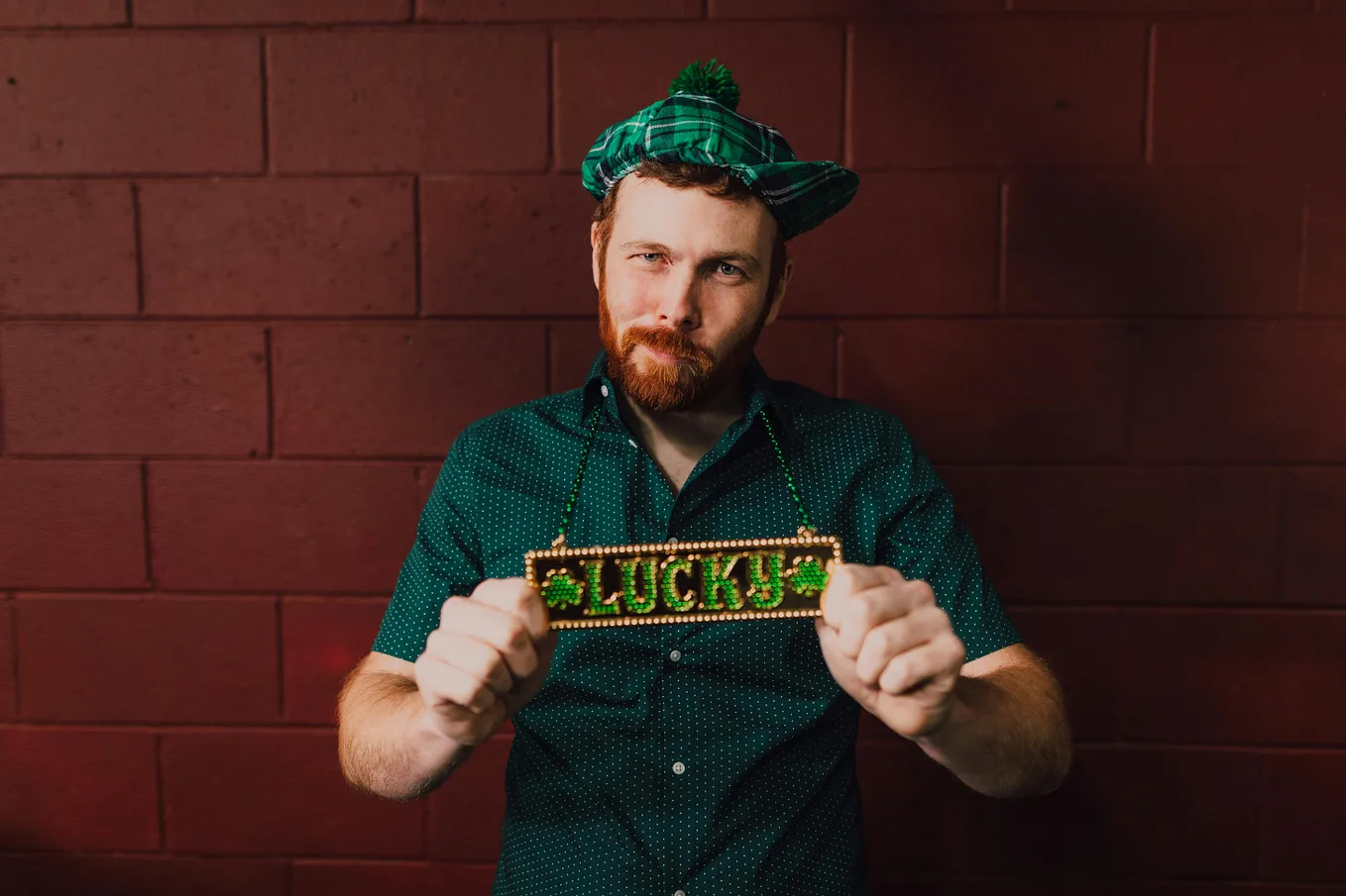 A red-haired man with a beard wearing a plaid green hat and green shirt holding a sign around his neck that says “Lucky” in sparkling green letters