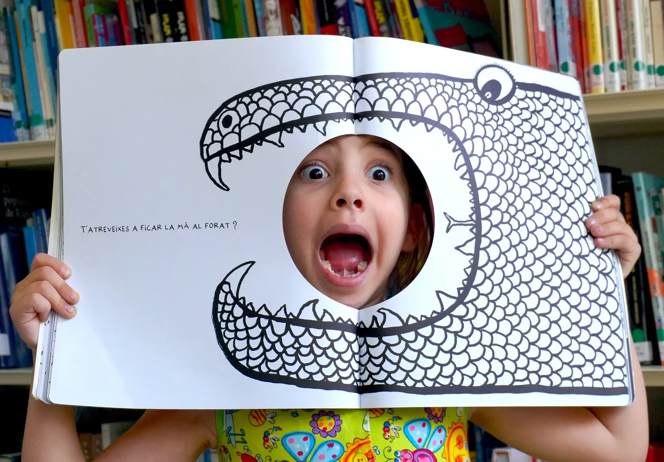 A young child holds up a book page exposing her face through a hole that puts her firmly in the mouth of a snake