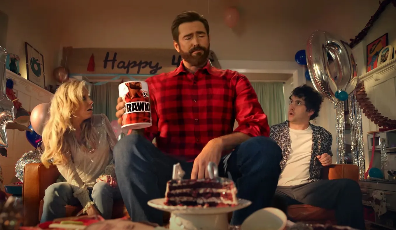The Brawny Man mascot holding Brawny paper towels sitting on a couch with two surprised people.