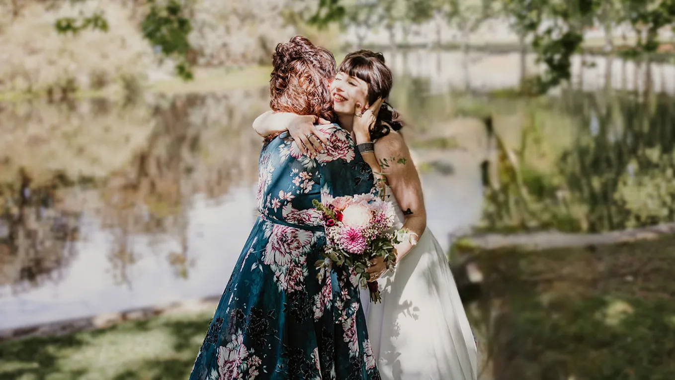 A smiling bride and woman in a floral dress hug, a picturesque lake in the background.