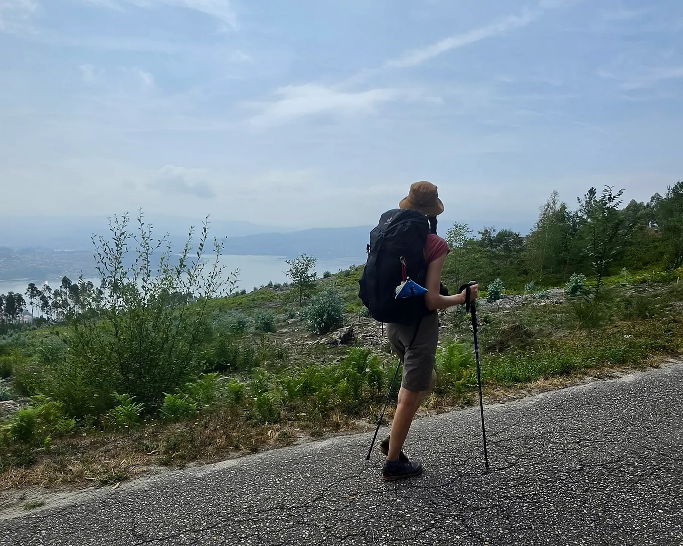 A woman, seen from behind, walks uphill using walking poles, a black pack on her back and wearing a beige bucket hat and knee-length shorts. There is grass next to the path, with the ocean visible in the distance.